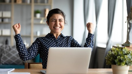 Junge Frau sitzt vor einen Computer und hält die Arme in freudiger Pose.
