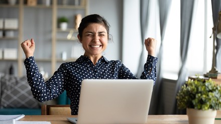 Junge Frau sitzt vor einen Computer und hält die Arme in freudiger Pose.