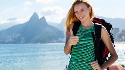 Junge Frau mit Rucksack steht lächelnd am Meer vor einer Bergkulisse und zeigt den Daumen nach oben.