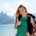 Junge Frau mit Rucksack steht lächelnd am Meer vor einer Bergkulisse und zeigt den Daumen nach oben.