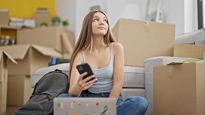Junge Frau sitzt zwischen Umzugskartons und schaut auf ihr Handy.