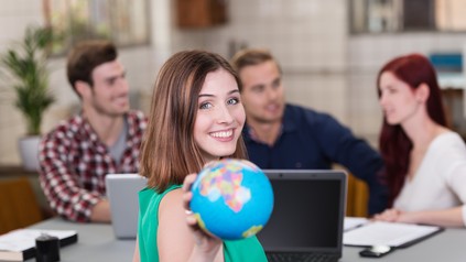 Junge Frau in grünem Oberteil hält lächelnd einen Globus in die Kamera. Im Hintergrund sitzen drei junge Erwachsene mit einem Laptop an einem Tisch.