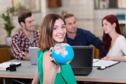Junge Frau in grünem Oberteil hält lächelnd einen Globus in die Kamera. Im Hintergrund sitzen drei junge Erwachsene mit einem Laptop an einem Tisch.