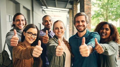 Weibliche und männliche Studierende auf Unigelände, die alle den Daumen hoch zeigen und in die Kamera lächeln.