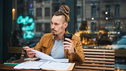 Student mit Dreadlocks sitzt mit Kaffee draußen im Cafè und lernt.