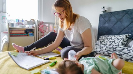 Junge Mutter, die auf dem Bett lernt und gleichzeitig ihr Baby mit der Flasche füttert.