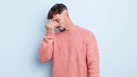 Junger Mann in rosa Pullover vor hellblauem Hintergrund, der vor Scham die Hand vor dem Gesicht hat.