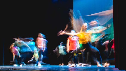 Performancekünstler:innen auf Bühne mit bunten Oberteilen und schwarzen Hosen.