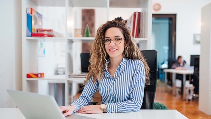 Junge Frau mit lockigen Haaren und Brille an ihrem Arbeitsplatz im Büro.