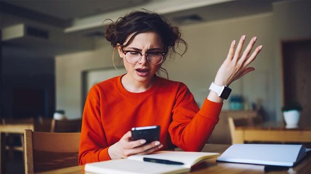 Genervte Studentin, die mit ihrem am Handy am Tisch sitzt.