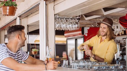 Barkeeper in Strandbar, der gerade etwas mixt und einem Kunden zulächelt, der auf einem Barhocker sitzt..