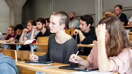 Studierende auf Holzbänken in Vorlesungssaal.