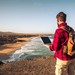 Mann in Karohemd an Strand mit Rucksack und Laptop.