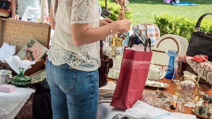 Frau auf Flohmarkt, vor einem Stand mit diversen Dingen wie Tellern, Handtaschen und Vasen.