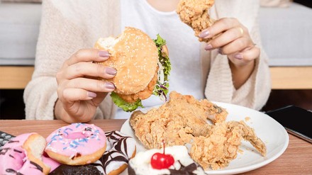 Essen ohne Sättigungsgefühl? (Foto: © stock.adobe.com/doucefleur) Viel Essen wie Burger und Donuts vor einer jungen Frau auf dem Tisch.