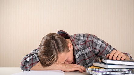 Müde vom Studieren oder Panik, das Studium nicht in der Regelstudienzeit zu beenden? (Foto: © Pixel-Shot / stock.adobe.com) Junger Student liegt mit dem Kopf auf dem Tisch. Neben ihm liegt ein Stapel mit Büchern.
