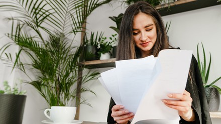 Bevor ihr den Vertrag unterzeichnet, solltet ihr ihn gut durchlesen. (Foto: © bodnarphoto/stock.adobe.com) Eine junge Frau sichtet wichtige Dokumente. Auf dem Tisch vor ihr steht ein zugeklappter Laptop und eine Kaffeetasse.