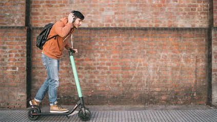Junger Mann mit Rucksack und Kopfhörern, der neben einem grünen City Roller auf dem Gehsteig geht.