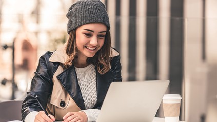 Eine junge Frau sitzt im Straßencafé mit aufgeklapptem Laptop und macht sich Notizen. Vor ihr auf dem Tisch steht ein Pappbecher.