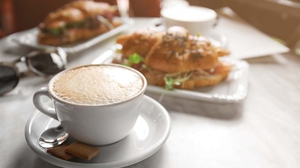 Nahaufnahme von voller Kaffeetasse und belegtem Sandwich auf einem Bistrotisch.