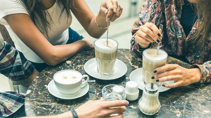Ausschnitt von rundem Tisch im Außenbereich eines Cafés mit verschiedenen Kaffeespezialitäten und den Armen und Oberkörpern von zwei Frauen und einem Mann.