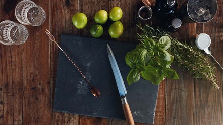 Schneidebrett mit Messer, Cocktaillöffel, Limetten und Minze auf Holztisch.