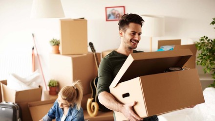 Zwei Studierende mit Umzungskartons in einer Wohnung. Sie sitzt am Boden, während er eine Kiste trägt.