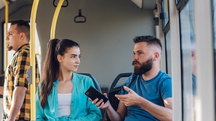 Ein bärtiger Mann sitzt zusammen mit einer jungen Frau in der Tram. Er hält ein Smartphone in der Hand.