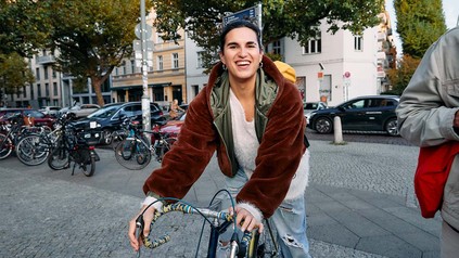 Lächelnde, junge Frau in Jeans und Jacke auf ihrem Fahrrad in der Stadt.