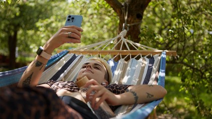 Diverse Person mit Handy in der Hand, die in Hängematte in der Natur liegt.