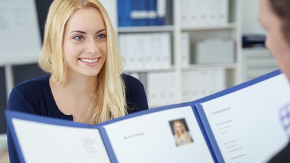 Eine junge Frau sitzt im Bewerbungsgespräch. Sie trägt ein schlichtes Outfit und lächelt den Personaler an, der in ihrer Bewerbungsmappe blättert.