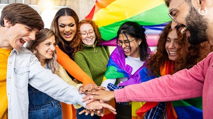 Das Bild zeigt verschiedene Personen unterschiedlichster Geschlechter. Im Hintergrund ist die Regenbogenflagge zu sehen.