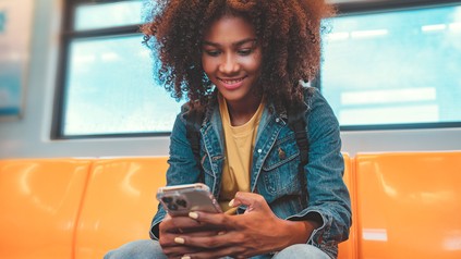 Eine junge Frau mit dunklerem Tein sitzt in der U-Bahnstation und schaut auf ihr Mobiltelefon. Sie lacht.
