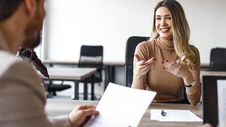 Eine junge Frau sitzt bei einem Mann in einem Vorstellungsgespräch. Die Frau lacht.