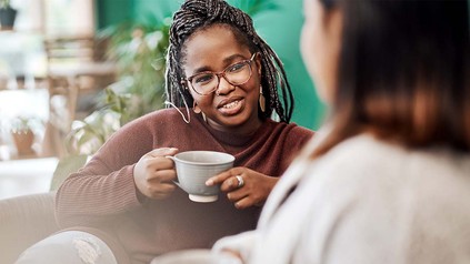 Junge Woman of Color mit Brille und Teetasse, die auf dem Sofa einer anderen Frau gegenübersitzt.