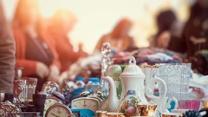 Nahaufnahme von Flohmarkttisch mit Teekannen und Co., dahinter Abenddämmerung und verschwommene Menschen.
