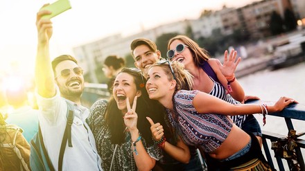 Studierendengruppe, die sommerlich gekleidet auf einer Brücke mit Sicht aufs Wasser ein Selfie macht.