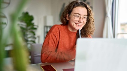 Junge Studentin mit Brille an Schreibtisch vor dem Laptop - lächelnd.