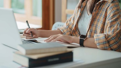 Schreibtisch mit Laptop und Büchern, davor Torso von Frau im Karohemd zu sehen.