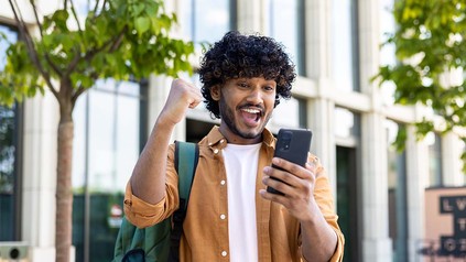 Junger Mann, der auf dem Unigelände auf sein Handy sieht und sich freut.
