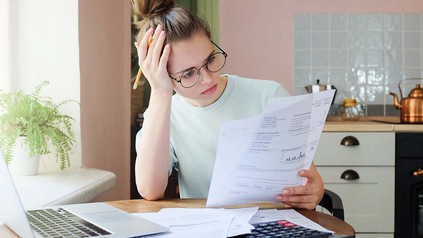 Junge Frau sitzt mit Laptop und Taschenrechner am Küchentisch und hat Finanzformulare in der Hand.