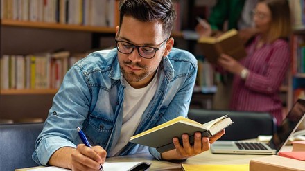 Junger Mann mit Brille, Laptop und Büchern an Arbeitsplatz in Unibibliothek.