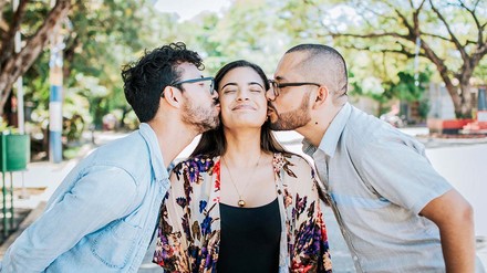 Zwei Männer, die der Frau in der Mitte von beiden Seiten einen Kuss auf die Wange drücken.