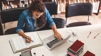 Junge Frau am Schreibtisch in Bibliothek mit Laptop, Büchern und Schreibmaterialien.