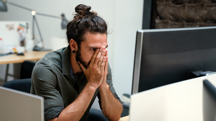 Junger Mann mit Dutt, der mit den Händen über dem Gesicht vor seinem PC im Büro sitzt.