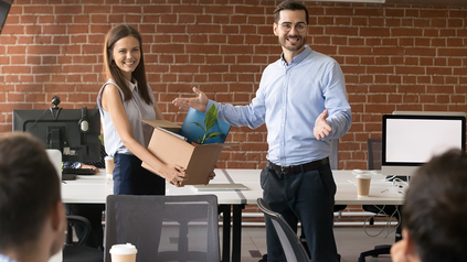 Junge Frau wird an ihrem ersten Arbeitstag im Büro den neuen Kolleg:innen vorgestellt - sie trägt einen Karton.