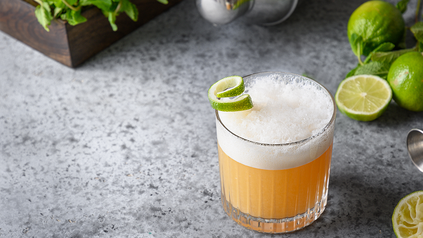 Whiskey Sour auf Arbeitsfläche mit Limetten und Cocktailshaker.
