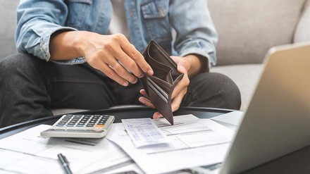 Torso von jungem Mann mit leeren Geldbeutel in der Hand – vor ihm Rechnungen und ein Taschenrechner.