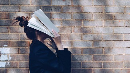 Junge Frau mit Dutt im Blazer vor Steinwand, schlägt sich Buch an den Kopf.