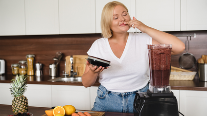 Junge Frau, die aus verschiedenem Obst und Gemüse einen Smoothie in der Küche kredenzt.